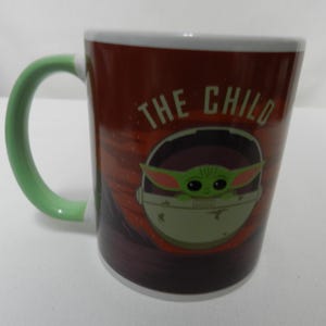 Op de afbeelding: Een keramische mok met een groene handgreep en witte rand, met "The Child" uit The Mandalorian. Het ontwerp van de mok bevat de tekst "THE CHILD" en een afbeelding van het personage in een pod, tegen een roodbruine achtergrond.