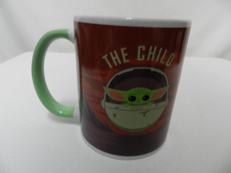 Op de afbeelding: Een keramische mok met een groene handgreep en witte rand, met "The Child" uit The Mandalorian. Het ontwerp van de mok bevat de tekst "THE CHILD" en een afbeelding van het personage in een pod, tegen een roodbruine achtergrond.