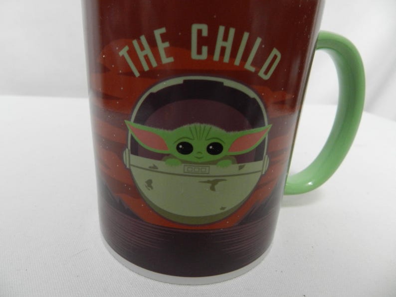 Op de afbeelding: Een keramische mok met het personage "The Child" uit de Star Wars-serie. De mok heeft een donkerrode achtergrond met de woorden "THE CHILD" in het wit. De handgreep is lichtgroen en de afbeelding van het personage staat in een lichtgroene drager.
