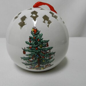 May include: White ceramic Christmas ornament with a festive tree design. The ornament has cut-out star shapes around the top and a red ribbon for hanging. The tree is decorated with colorful ornaments.