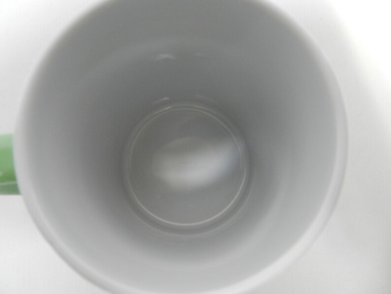 Op de afbeelding: Een witte keramische mok met een lichtgroen handvat. De binnenkant van de mok is lichtgrijs van kleur. De mok is leeg en lijkt schoon te zijn.