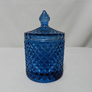 Könnte beinhalten: Ein kobaltblaues Glasgefäß mit Rautenmuster und passendem Deckel. Das Gefäß hat eine runde Form und eine dekorative Spitze oben. Das Gefäß steht auf einer weißen Oberfläche.