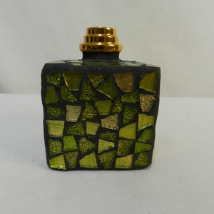 May include: A square glass bottle featuring a mosaic exterior composed of various shades of green and gold glass shards set in dark grout. The bottle is topped with a tiered, metallic gold cap that has a stepped, cylindrical design.