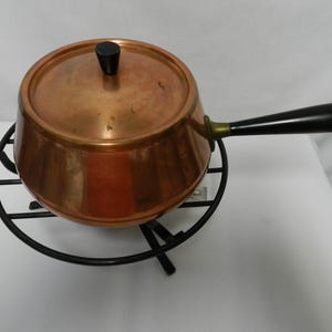 May include: A copper saucepan with a black handle and lid, resting on a black metal trivet. The pot has a shiny, reflective surface. The handle is angled, and the lid has a black knob. A vintage kitchenware item.