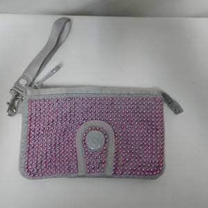 May include: Gray wristlet wallet covered in pink rhinestones. The wallet has a silver zipper, a wrist strap, and a silver clasp. The front of the wallet has a decorative silver button.