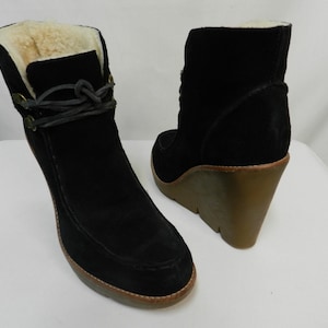 May include: A pair of black suede ankle boots featuring a thick, tan wedge heel and a cream-colored shearling lining. The boots include a dark gray lace-up detail around the ankle, secured with small metal eyelets and a knot at the front.