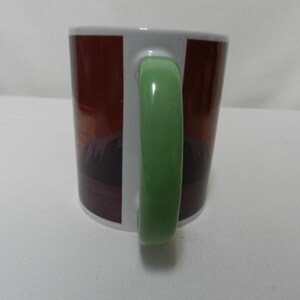 Op de afbeelding: Een witte keramische mok met een groene handgreep. De mok heeft een bruin en donkerrood ontwerp aan de buitenkant. De mok staat op een wit oppervlak.