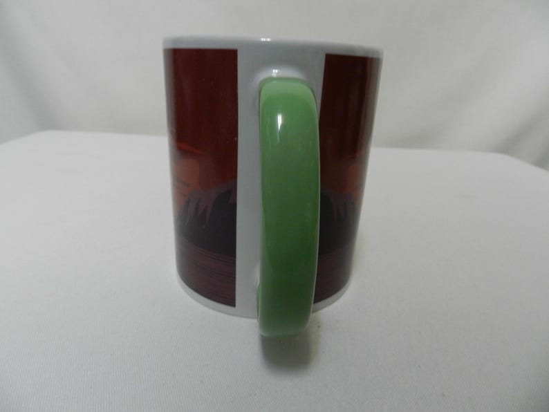 Op de afbeelding: Een witte keramische mok met een groene handgreep. De mok heeft een bruin en donkerrood ontwerp aan de buitenkant. De mok staat op een wit oppervlak.