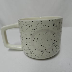 May include: Cream-colored ceramic mug with black speckles and a square handle. The mug has a cylindrical shape and a light-colored interior. The mug is suitable for coffee, tea, or other beverages.