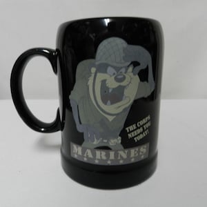 May include: Black ceramic mug featuring a cartoon Taz character in a military uniform. The mug has the words "MARINES" and "THE CORPS NEEDS YOU TODAY!" printed on it. The mug has a black handle and a glossy finish.