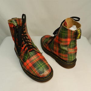 May include: A pair of ankle-high boots featuring a red and green plaid tartan pattern. The boots have black laces, black soles, and a yellow heel pull tab with the text AirWair printed in black. The design includes a rounded toe and eyelets.
