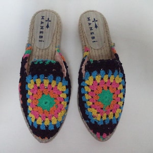 May include: A pair of colorful crochet espadrilles with a black background and a multi-colored granny square design. The espadrilles have a jute sole and the brand name "MANEBI" is printed on the top of each shoe.