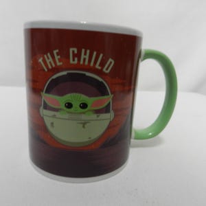 Op de afbeelding: Een keramische mok met het personage "The Child" uit The Mandalorian. De mok heeft een rode en bruine achtergrond met het personage in een pod. De handgreep is groen en de rand is wit.