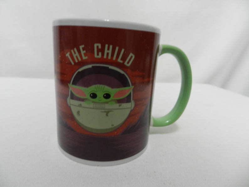 Op de afbeelding: Een keramische mok met het personage "The Child" uit The Mandalorian. De mok heeft een rode en bruine achtergrond met het personage in een pod. De handgreep is groen en de rand is wit.