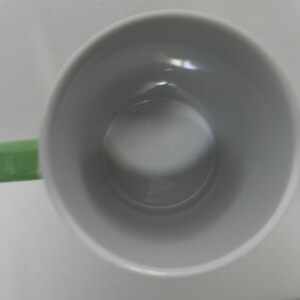 Op de afbeelding: Een witte keramische mok met een lichtgroen oor. De binnenkant van de mok is lichtgrijs van kleur. De mok is leeg en van bovenaf gezien.