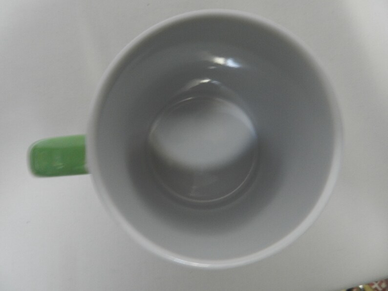 Op de afbeelding: Een witte keramische mok met een lichtgroen oor. De binnenkant van de mok is lichtgrijs van kleur. De mok is leeg en van bovenaf gezien.