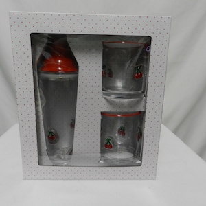 May include: A cocktail shaker set in a white box with red polka dots. The set includes a clear cocktail shaker with a red top and two clear glasses, all decorated with cherry designs. The glasses have red rims.