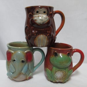 May include: Three ceramic mugs with animal faces. The mugs are brown, blue, and red with green accents. The first mug features a monkey face, the second mug features an elephant face, and the third mug features a frog face.