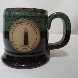 Op de afbeelding: Een keramische mok met een donkergroen en bruin glazuur. De mok heeft een cirkelvormig embleem met een vuurtorenillustratie en de tekst "Lighthouse Bakery, Dauphin Island, Alabama."
