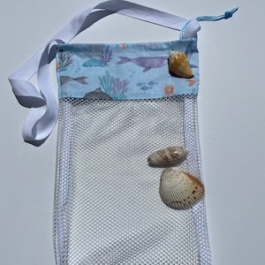 May include: White mesh bag with a blue and white fabric top featuring a sea life print. The bag has a white drawstring and a white strap. There are three seashells inside the bag.