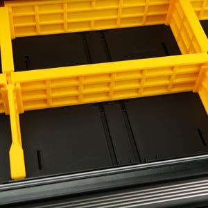 May include: Close-up of a black storage container with bright yellow dividers. The dividers are rectangular with a grid-like pattern, creating multiple compartments. The container appears to be made of durable plastic, designed for organization.
