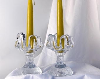 May include: Two clear glass candle holders with yellow candles. The candle holders have a decorative, scalloped design around the top. The candles are a bright yellow color and are unlit. The background is a white cloth.