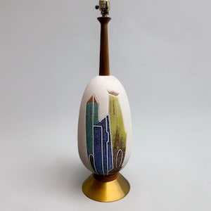 May include: A table lamp with a tall, egg-shaped white ceramic base featuring abstract blue, green, and brown painted designs. The lamp has a slender dark wood neck, a metallic gold-toned conical base, and a silver-colored metal light socket.
