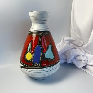 May include: A decorative ceramic vase with a red body and a white neck. The vase features a painted design of fruits and bottles in blue, yellow, and red. The vase has a round base and a narrow neck.