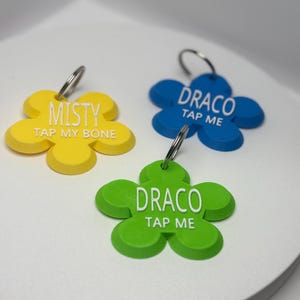 以下が含まれることがあります： 黄色、青、緑の3つの花型のペットタグ。黄色のタグには「MISTY TAP MY BONE」と書かれています。青と緑のタグには「DRACO TAP ME」と書かれています。各タグにはシルバーのキーリングが付いています。