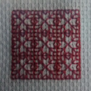 May include: A red embroidered design on a gray fabric background. The design is a square with a repeating pattern of small red shapes.