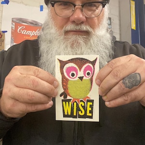 May include: A sticker with a colourful owl illustration. The owl has pink eyes and a yellow body. The word "WISE" is printed in yellow on a black rectangle below the owl. The owl design is made of small dots.