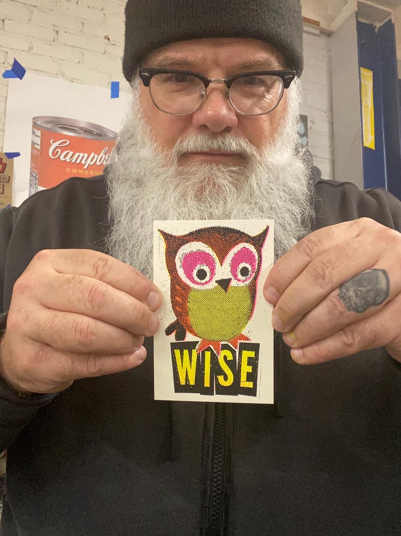 May include: A sticker with a colourful owl illustration. The owl has pink eyes and a yellow body. The word "WISE" is printed in yellow on a black rectangle below the owl. The owl design is made of small dots.