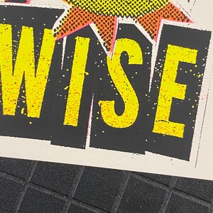 May include: Close-up of a print with the word "WISE" in bright yellow, outlined in pink, against a black background. The top of the print features a yellow and orange burst design. The background is white.