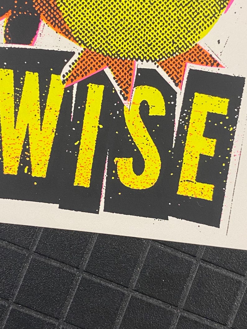 May include: Close-up of a print with the word "WISE" in bright yellow, outlined in pink, against a black background. The top of the print features a yellow and orange burst design. The background is white.