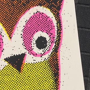 May include: Close-up of a pop art style owl illustration. The owl's eye is pink with a black pupil. The owl's feathers are yellow and orange, with black accents. The background is white with black speckles.