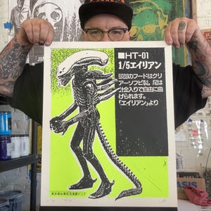 以下が含まれることがあります： 黒とネオングリーンのポスター。エイリアンの詳細なイラストが描かれています。ポスターには日本語のテキストと「HT-01 1/5 エイリアン」というタイトルが含まれています。アートワークは「LA HONDA」と書かれた黒い帽子をかぶった人が持っています。