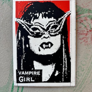 May include: A rectangular sticker featuring a high-contrast black and white graphic of a person with long hair, wearing pointed cat-eye glasses and displaying sharp fangs. The background is red, and the text VAMPIRE GIRL is printed in white.