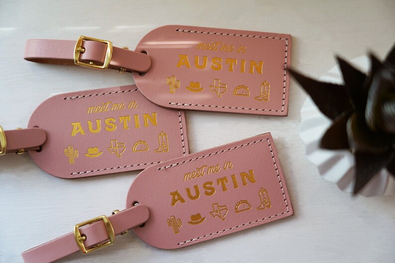 K&ouml;nnte beinhalten: Drei rosafarbene Leder-Gep&auml;ckanh&auml;nger mit goldener Aufschrift "Meet me in Austin". Jeder Anh&auml;nger ist mit einem goldenen Kaktus-, Cowboyhut-, Texasstern-, Taco- und Cowboystiefel-Symbol verziert.