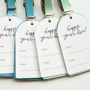 May include: Five luggage tags with white card stock and black calligraphy that reads "happy you're here!" Each tag has a blank space for a name and table number. The tags are attached to leather straps in various colors: pink, blue, turquoise, green, and mint.