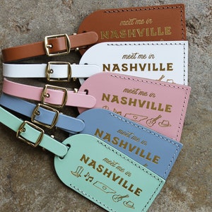 May include: Five leather luggage tags with gold buckles and the text "meet me in NASHVILLE" in gold lettering. The tags are in different colors: brown, white, pink, blue, and light green. The blue tag has a small illustration of a guitar, a cowboy hat, and the state of Tennessee.