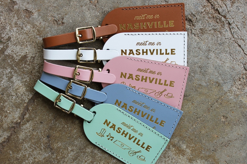 May include: Five leather luggage tags with gold buckles and the text "meet me in NASHVILLE" in gold lettering. The tags are in different colors: brown, white, pink, blue, and light green. The blue tag has a small illustration of a guitar, a cowboy hat, and the state of Tennessee.