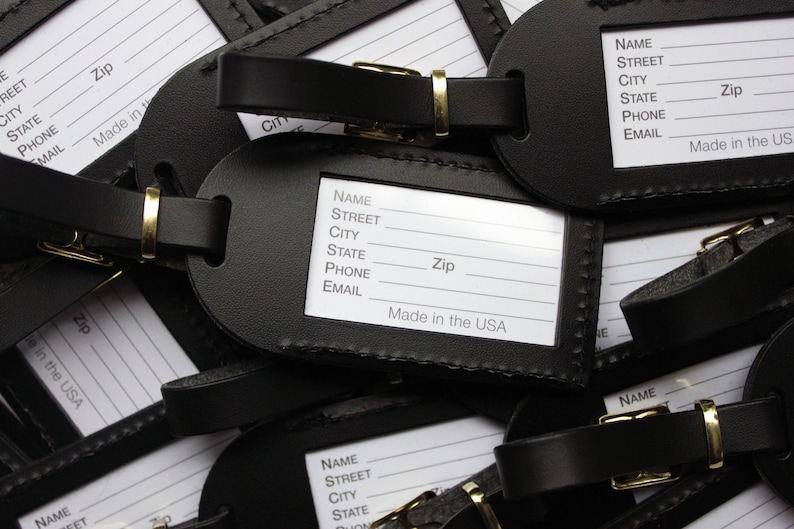 May include: A close-up of a stack of black leather luggage tags with white labels. The labels have fields for name, street, city, state, zip, phone, and email. The tags are made in the USA.