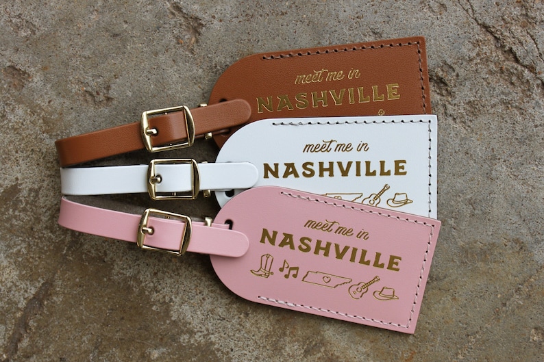 May include: Three leather luggage tags with gold lettering that reads "meet me in NASHVILLE". The tags are brown, white, and pink. The pink tag has a small design of a boot, musical notes, the state of Tennessee, a guitar, and a cowboy hat.