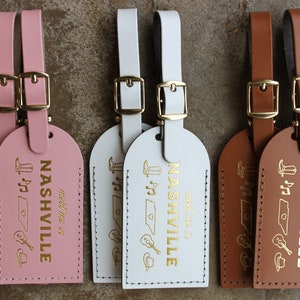 May include: Three leather luggage tags with gold accents. Each tag features the word "Nashville" with the text "meet me in" below. The tags are pink, white, and brown.