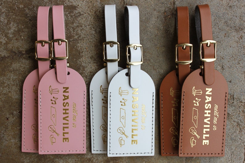 May include: Three leather luggage tags with gold accents. Each tag features the word "Nashville" with the text "meet me in" below. The tags are pink, white, and brown.