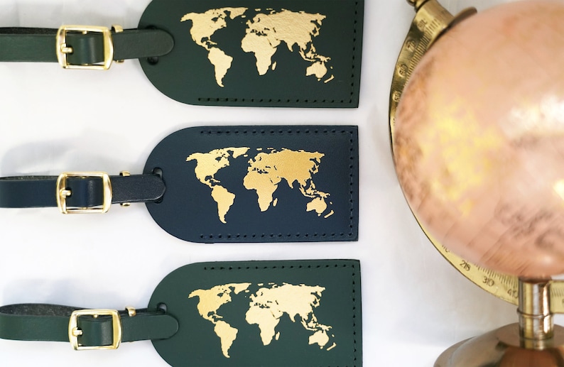 Op de afbeelding: Drie leren bagagelabeltjes met gouden wereldkaarten van folie. De labeltjes zijn groen, donkerblauw en donkergroen. Elk labeltje heeft een gouden metalen gesp en een bandje.