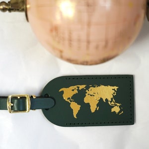 Op de afbeelding: Een donkergroen leren bagagelabel met een gouden wereldkaartontwerp. Het label heeft een messing gesp en een band.