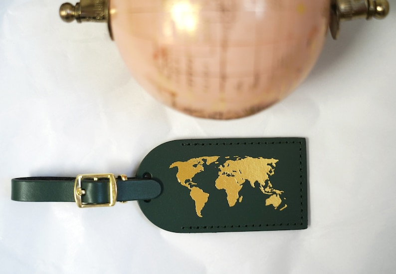 Op de afbeelding: Een donkergroen leren bagagelabel met een gouden wereldkaartontwerp. Het label heeft een messing gesp en een band.