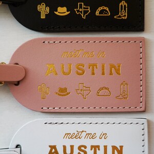 K&ouml;nnte beinhalten: Drei Leder-Gep&auml;ckanh&auml;nger mit goldenen N&auml;hten und goldenen Buchstaben. Der obere Anh&auml;nger ist schwarz mit goldenen Symbolen eines Kaktus, eines Cowboyhuts, einer Umrisskarte von Texas, eines Tacos und eines Cowboystiefels. Der mittlere Anh&auml;nger ist rosa mit den gleichen Symbolen und dem Text "meet me in AUSTIN". Der untere Anh&auml;nger ist wei&szlig; mit den gleichen Symbolen und dem Text "meet me in AUSTIN".