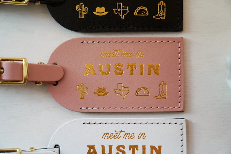 K&ouml;nnte beinhalten: Drei Leder-Gep&auml;ckanh&auml;nger mit goldenen N&auml;hten und goldenen Buchstaben. Der obere Anh&auml;nger ist schwarz mit goldenen Symbolen eines Kaktus, eines Cowboyhuts, einer Umrisskarte von Texas, eines Tacos und eines Cowboystiefels. Der mittlere Anh&auml;nger ist rosa mit den gleichen Symbolen und dem Text "meet me in AUSTIN". Der untere Anh&auml;nger ist wei&szlig; mit den gleichen Symbolen und dem Text "meet me in AUSTIN".
